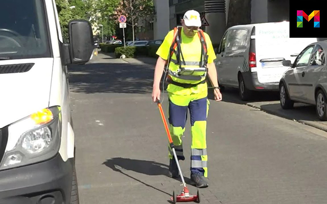 Ein Mann mit Warnkleidung geht mit einem Gasrohrnetzüberprüfungsgeraät über eine Straße